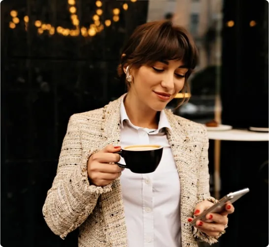 Frau mit Kaffee und Smartphone vor einem Café, stilvoll gekleidet.